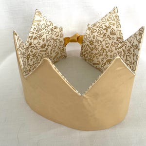 May include: A crown with a solid matte gold exterior and a white interior lining featuring a repeating gold pattern of holiday icons including reindeer, bells, gifts, and candy canes. The crown is secured at the back with a small gold ribbon.