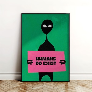 Op de afbeelding: Een ingelijste kunstdruk met een zwart alienfiguur tegen een groene achtergrond. De alien houdt een roze bord vast met de zwarte tekst "HUMANS DO EXIST". Het kunstwerk wordt op een houten vloer getoond.