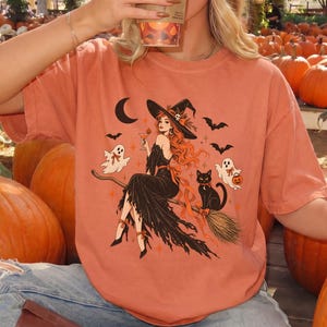 May include: A burnt orange t-shirt featuring a graphic print of a witch with long red hair sitting on a broomstick, accompanied by a black cat, two ghosts, bats, and a crescent moon. The shirt is worn with blue jeans and brown boots.