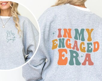 May include: A light gray sweatshirt with the text "In My Engaged Era" in retro colors on the back. The front of the sweatshirt has the text "Mrs. Smith" in a cursive font. The sweatshirt is a sports gray color.