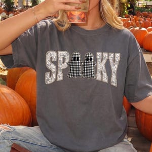 May include: A charcoal gray short-sleeve t-shirt featuring the word SPOOKY in white floral patterned letters, with two black and white gingham ghosts replacing the letter O. The shirt is worn with light blue denim jeans and brown boots.