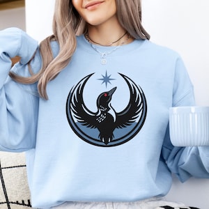 May include: Light blue sweatshirt featuring a black and gray graphic of a bird with outstretched wings, a red eye, and a star above it. The sweatshirt has long sleeves and a crew neck. The words "Light Blue" are in the upper left corner.