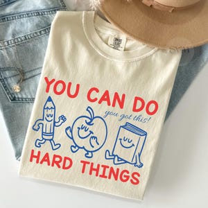 May include: An ivory t-shirt featuring red text that reads YOU CAN DO HARD THINGS and blue text that reads you got this! The graphic includes blue line art of a smiling pencil, an apple, and a book. Also shown are blue jeans and a tan hat.