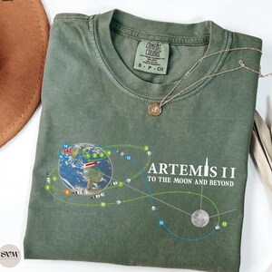 May include: Moss green t-shirt with a graphic of Earth, a rocket, and the moon, with the text "ARTEMIS II TO THE MOON AND BEYOND." The shirt has a crew neck and short sleeves. A silver necklace with a pendant is draped on the shirt.