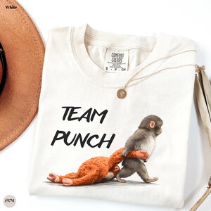 May include: White t-shirt with the words "TEAM PUNCH" in black. The shirt features a graphic of two monkeys, one orange and one gray, in a playful pose. A gold necklace with a pendant is draped across the shirt.