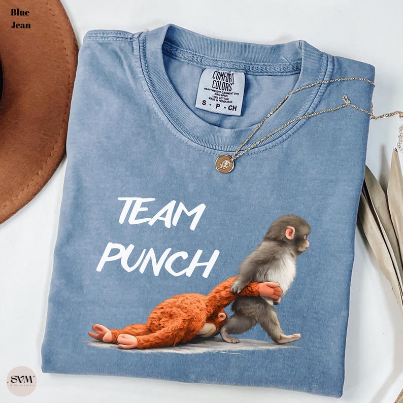 May include: Blue jean Comfort Colors t-shirt with the text "TEAM PUNCH" in white. The shirt features a graphic of a monkey carrying an orange stuffed animal. A gold necklace with a pendant is also visible.