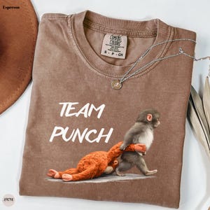 May include: Espresso-colored t-shirt with the words "TEAM PUNCH" in white. The graphic depicts a monkey carrying another monkey. The shirt has a crew neck and a Comfort Colors tag. A silver necklace with a pendant is draped across the shirt.