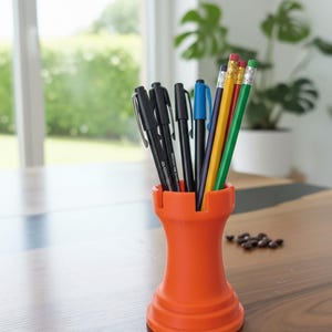 May include: An orange pen holder, resembling a castle turret, containing black pens, a blue pen, and pencils in yellow, red, and green. It rests on a wooden table. The background shows a window and plants, out of focus.