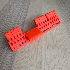 Op de afbeelding: Fel oranje plastic gereedschap met een reeks sleuven en cijfers erop gedrukt. De cijfers 1 tot en met 20 zijn zichtbaar, samen met een reeks sleuven. De tool is ontworpen om objecten vast te houden en te meten.