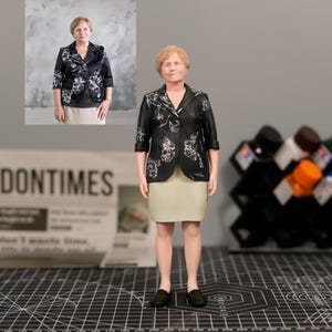 Könnte beinhalten: 3D-gedruckte Figur einer Frau in einer schwarzen Jacke mit weißen Blumenmustern, einem beigen Rock und schwarzen Schuhen. Die Figur steht auf einer schwarzen Schneidematte. Eine Zeitung mit "LONDONTIMES" ist im Hintergrund.