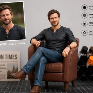 May include: A custom figurine of a man in a dark gray Henley shirt and blue jeans, seated in a brown leather armchair. The figurine holds a black mug and wears a watch. A framed photo and a newspaper are in the background.