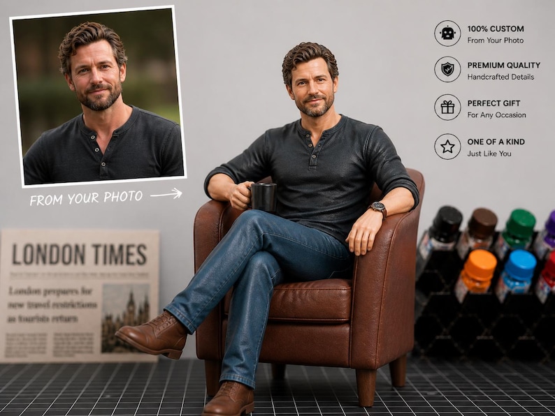 May include: A custom figurine of a man in a dark gray Henley shirt and blue jeans, seated in a brown leather armchair. The figurine holds a black mug and wears a watch. A framed photo and a newspaper are in the background.
