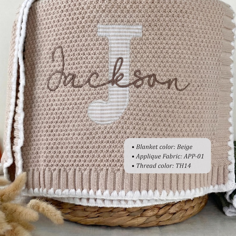 May include: A beige knit blanket featuring a large, striped letter J applique and the name Jackson embroidered in brown script. The blanket has a white crocheted border and a label with the text: Blanket color: Beige, Applique Fabric: APP-01, Thread color: TH14.