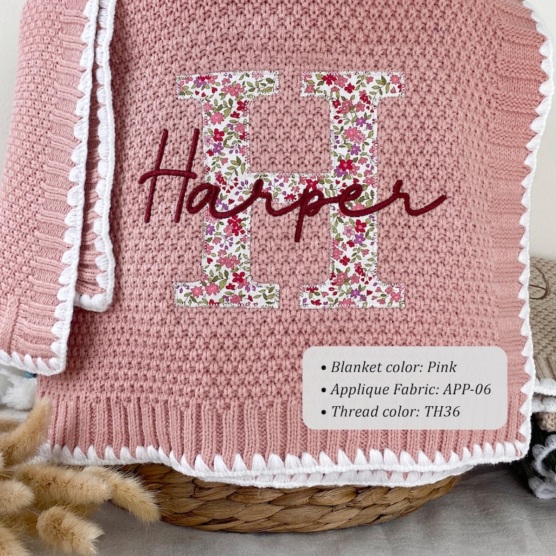 May include: A pink knitted blanket featuring a large letter H applique in a floral pattern with the name Harper embroidered in dark red script across the center. The blanket has a white crocheted border and a label listing Blanket color: Pink, Applique Fabric: APP-06, and Thread color: TH36.