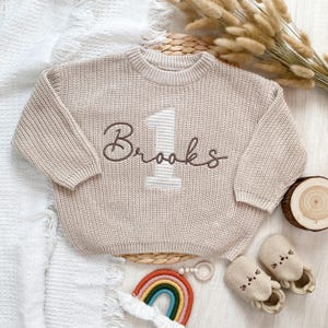 May include: A beige knit sweater with the name "Brooks" embroidered in brown script and a number "1" with a striped pattern. The sweater is part of a baby clothing set, with matching shoes and a rainbow toy.