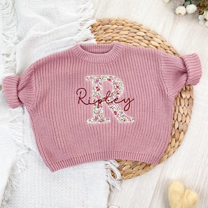 Może przedstawiać: Długi sweter z dzianiny w kolorze przybrudzonego r&oacute;żu z dużą aplikacją litery R w kwiatowy wz&oacute;r na środku. Imię Ripley jest wyhaftowane ciemnoczerwoną kursywą na literze. Sweter posiada prążkowany dekolt w kształcie okrągłym.