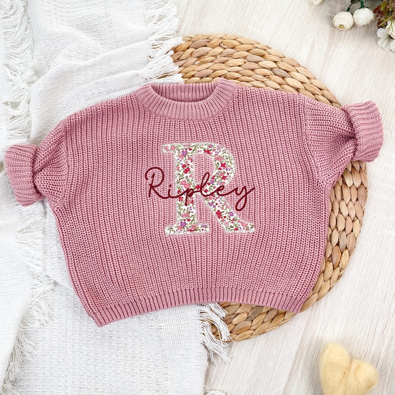 Może przedstawiać: Długi sweter z dzianiny w kolorze przybrudzonego r&oacute;żu z dużą aplikacją litery R w kwiatowy wz&oacute;r na środku. Imię Ripley jest wyhaftowane ciemnoczerwoną kursywą na literze. Sweter posiada prążkowany dekolt w kształcie okrągłym.