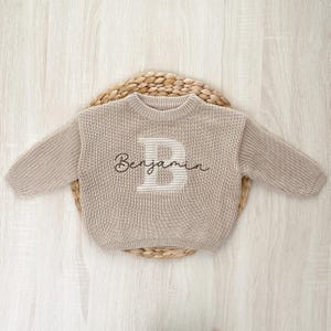 May include: A beige knit sweater featuring a large appliqué letter “B” in a striped design and the name “Benjamin” embroidered on the front, displayed on a woven circular mat.