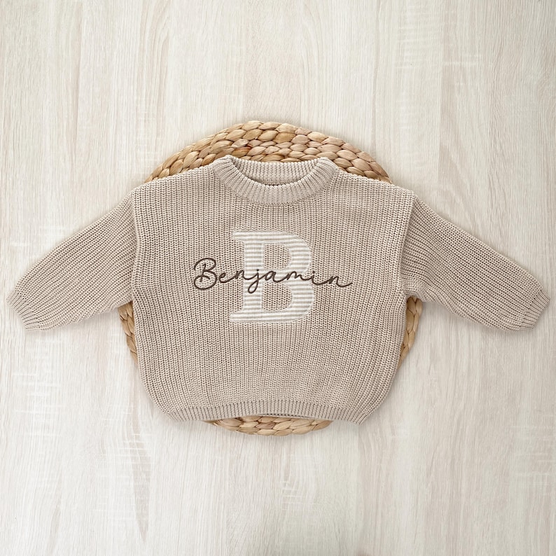 May include: A beige knit sweater featuring a large appliqué letter “B” in a striped design and the name “Benjamin” embroidered on the front, displayed on a woven circular mat.