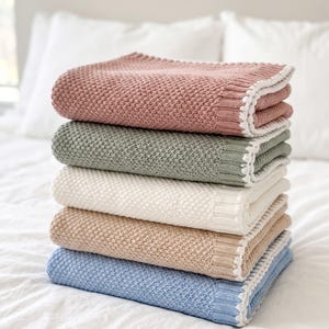 May include: A stack of five folded knit blankets in dusty rose, sage green, cream, beige, and light blue. Each blanket features a textured honeycomb stitch pattern, a ribbed border, and a decorative white scalloped edge along the folded side.