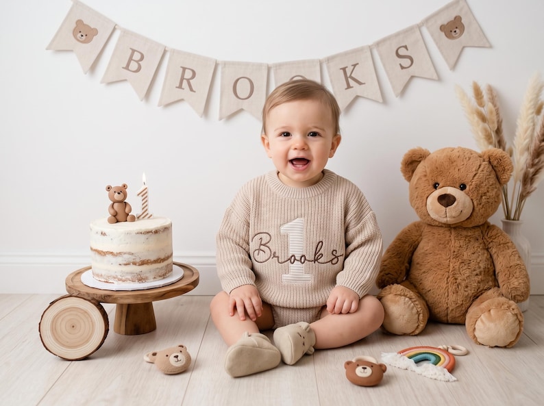Pode incluir: Um su&eacute;ter de malha bege com o n&uacute;mero 1 e o nome Brooks bordados na frente. O su&eacute;ter &eacute; usado por uma crian&ccedil;a sentada perto de um bolo com uma vela do n&uacute;mero 1, um urso de pel&uacute;cia e uma faixa com o nome Brooks e &iacute;cones de ursos.