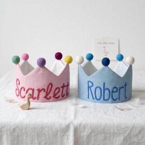 May include: Two fabric crowns with pom-poms on the points. The pink crown is embroidered with the name Scarlett in red, and the blue crown is embroidered with the name Robert in blue. A small wooden goose figure and a card labeled One Silly Goose.