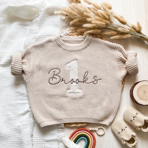 May include: A beige knit sweater with the number "1" applique embroidered and the name "Brooks" embroidered in brown. The sweater is surrounded by baby accessories, including shoes, a rainbow, and a wooden toy.