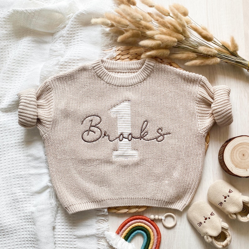 May include: A beige knit sweater with the number "1" applique embroidered and the name "Brooks" embroidered in brown. The sweater is surrounded by baby accessories, including shoes, a rainbow, and a wooden toy.