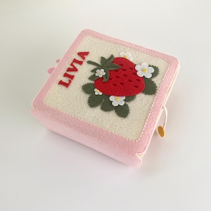 May include: A square, light pink felt box featuring a cream-colored top panel decorated with a red strawberry appliqué, dark green leaves, and small white flowers. The name LIVIA is stitched in red letters on the left side of the top panel.