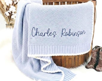 Pode incluir: Um cobertor de bebê de malha azul claro com o nome "Charles Robinson" bordado em azul marinho. O cobertor tem uma borda branca e está sobre uma cesta de madeira. Botas de cowboy marrons são visíveis.