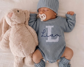 May include: A baby wearing a dusty blue ribbed knit long-sleeve romper with a white and blue gingham letter L and the name Liam embroidered in dark blue script, a matching pom-pom beanie, and booties, lying next to a plush beige stuffed rabbit.