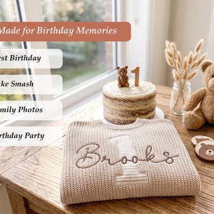 Pode incluir: Uma camisola de malha bege com o nome Brooks bordado e um n&uacute;mero um grande. A camisola &eacute; exibida ao lado de um bolo de anivers&aacute;rio com uma figura de urso e uma vela de n&uacute;mero um, um urso de peluche castanho e bases para copos de madeira.