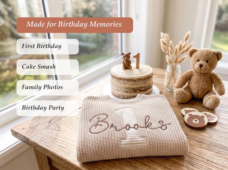 Pode incluir: Uma camisola de malha bege com o nome Brooks bordado e um n&uacute;mero um grande. A camisola &eacute; exibida ao lado de um bolo de anivers&aacute;rio com uma figura de urso e uma vela de n&uacute;mero um, um urso de peluche castanho e bases para copos de madeira.