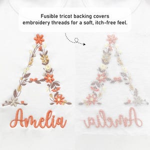 May include: White fabric featuring embroidered floral letter "A" and the name "Amelia" in orange thread. The text "Fusible tricot backing covers embroidery threads for a soft, itch-free feel" is also visible.