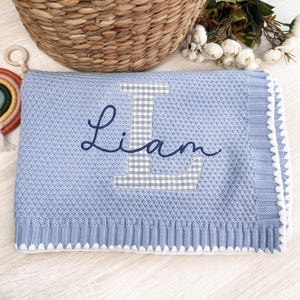 May include: A light blue knitted blanket featuring a textured pattern and a white scalloped edge. The center displays a large grey and white gingham letter L with the name Liam embroidered in dark blue cursive script across the letter.