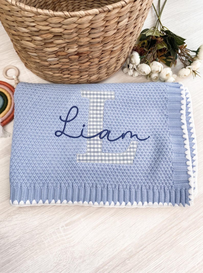 May include: A light blue knitted baby blanket with the name "Liam" embroidered in dark blue script. The letter "L" is a applique embroidered gingham pattern. The blanket has a white scalloped edge and a textured knit design. newborn boy blanket