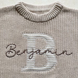 May include: A beige knitted sweater with a ribbed crew neck. The sweater has a large letter "B" with a striped pattern and the name "Benjamin" embroidered in a dark brown script.