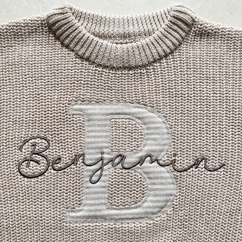 May include: A beige knitted sweater with a ribbed crew neck. The sweater has a large letter "B" with a striped pattern and the name "Benjamin" embroidered in a dark brown script.