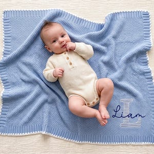 May include: A light blue knitted baby blanket with a white border and a textured pattern. The blanket features the name Liam embroidered in dark blue script over a large, light blue and white checkered letter L appliqu&eacute; in the bottom corner.