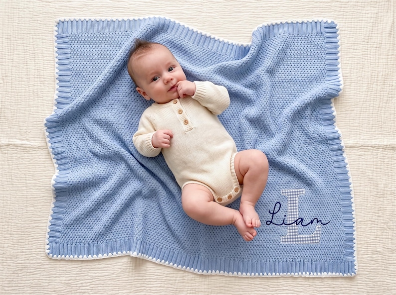 May include: A light blue knitted baby blanket with a white border and a textured pattern. The blanket features the name Liam embroidered in dark blue script over a large, light blue and white checkered letter L appliqu&eacute; in the bottom corner.