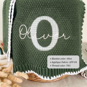 May include: An olive green knit blanket featuring a white scalloped edge and a large, central letter O in a white and grey gingham pattern. The name Oliver is embroidered in white cursive script over the letter. Includes text: Blanket color: Olive, Applique Fabric: APP-04, Thread color: TH1.