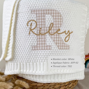 May include: A white knitted blanket featuring a large beige and white gingham letter R applique with the name Riley embroidered in gold script thread across the center. A small rectangular label lists Blanket color: White, Applique Fabric: APP-02, Thread color: TH2.