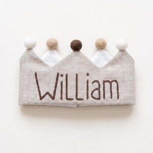 Pode incluir: Uma coroa de linho bege com o nome William bordado em fio castanho escuro. A coroa tem cinco pontas, cada uma rematada com uma pequena bola de feltro em cores alternadas branco, bege e castanho, criando um aspeto texturado e neutro.