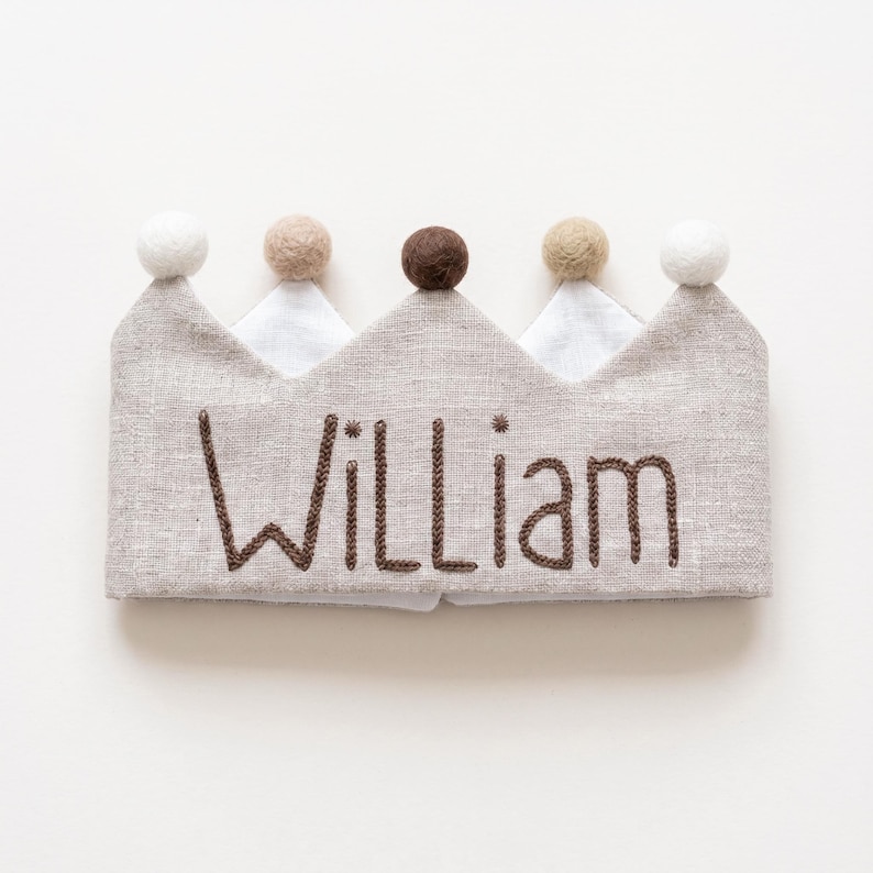 Pode incluir: Uma coroa de linho bege com o nome William bordado em fio castanho escuro. A coroa tem cinco pontas, cada uma rematada com uma pequena bola de feltro em cores alternadas branco, bege e castanho, criando um aspeto texturado e neutro.