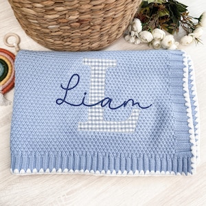 May include: A light blue knitted baby blanket with the name "Liam" embroidered in dark blue script. The letter "L" is a gingham pattern. The blanket has a white scalloped edge and a textured knit design. A wicker basket and flowers are in the background.