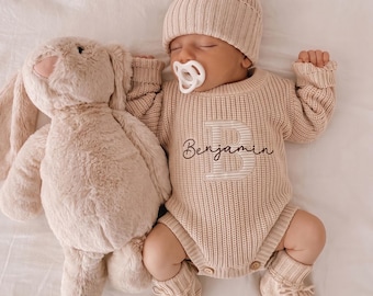 May include: A beige knitted long-sleeve romper featuring a large letter B with the name Benjamin embroidered in dark brown script. The outfit includes a matching ribbed beanie with a pom-pom and knitted booties, paired with a plush beige rabbit toy.
