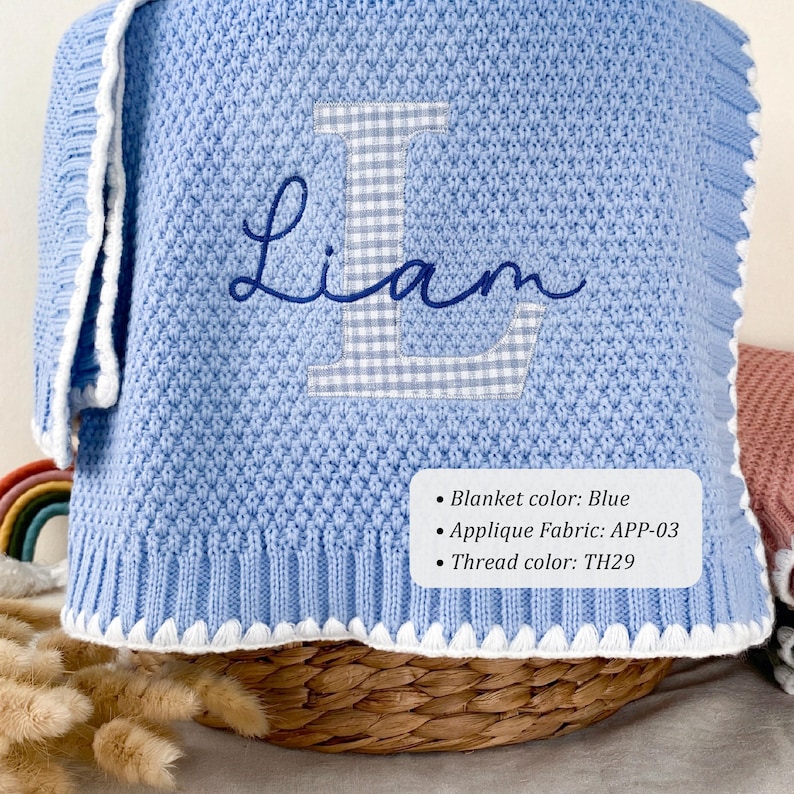 May include: A light blue knitted blanket featuring a white scalloped edge and a large, grey and white gingham letter L applique. The name Liam is embroidered in dark blue cursive script over the letter. Text on the blanket reads: Blanket color: Blue, Applique Fabric: APP-03, Thread color: TH29.
