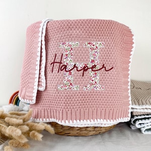 May include: A dusty pink knitted blanket featuring a white crocheted border. The centre displays a large letter H filled with a colourful floral pattern, with the name Harper embroidered in dark red cursive script across the letter.