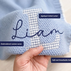 May include: A light blue knit blanket featuring a large white and blue gingham appliqu&eacute;d initial letter L with the name Liam embroidered in dark blue cursive script. The blanket is made of a textured knit yarn and includes labels pointing to the Appliqu&eacute; Initial Letter, Embroidered custom name, and Soft and breathable knit yarn.