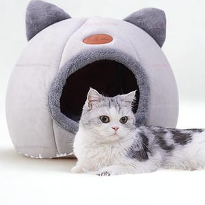 May include: A gray and white cat lounges beside a gray cat bed. The cat bed is shaped like a cat's head, with gray ears and a circular opening. The bed has a soft, plush appearance, designed for comfort.